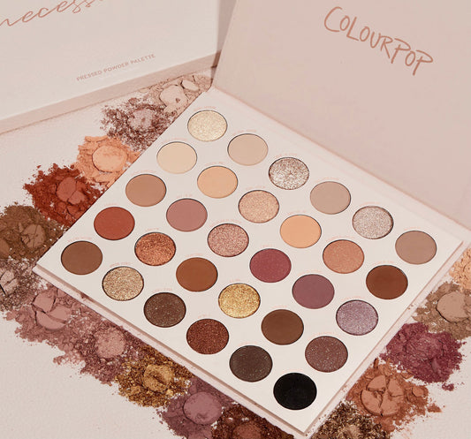 ColourPop Shadow Palette Bare Necessities lies open, showing 30 pressed neutral shades - matte and shimmer - ranging from ivory, taupe, and peach to bronze, plum, and black, arranged in rows amid scattered crushed pigments on a textured surface.