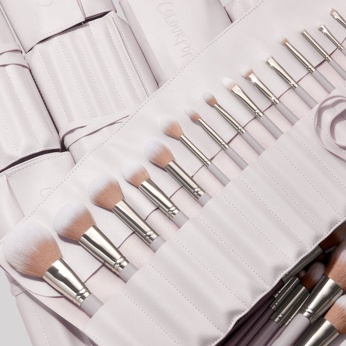 The Ultimate Stunner, a ColourPop Shadow Palette & Brush Set, displays white-tipped face and eye brushes with silver ferrules neatly lined in a pale stone faux-leather roll case, pastel handles visible and ready for precise application.