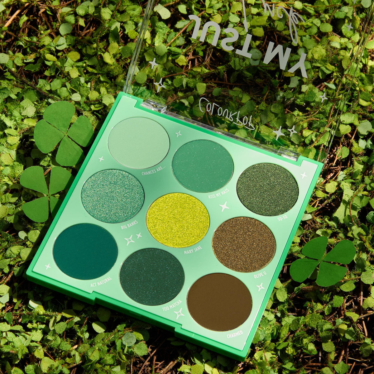 Just My Luck ColourPop Shadow Palette displays nine green eyeshadows - mint, lime, olive, and emerald - in matte and shimmer finishes, opened with a clear lid and star accents, set on a bed of clover for a luck-themed vibe.