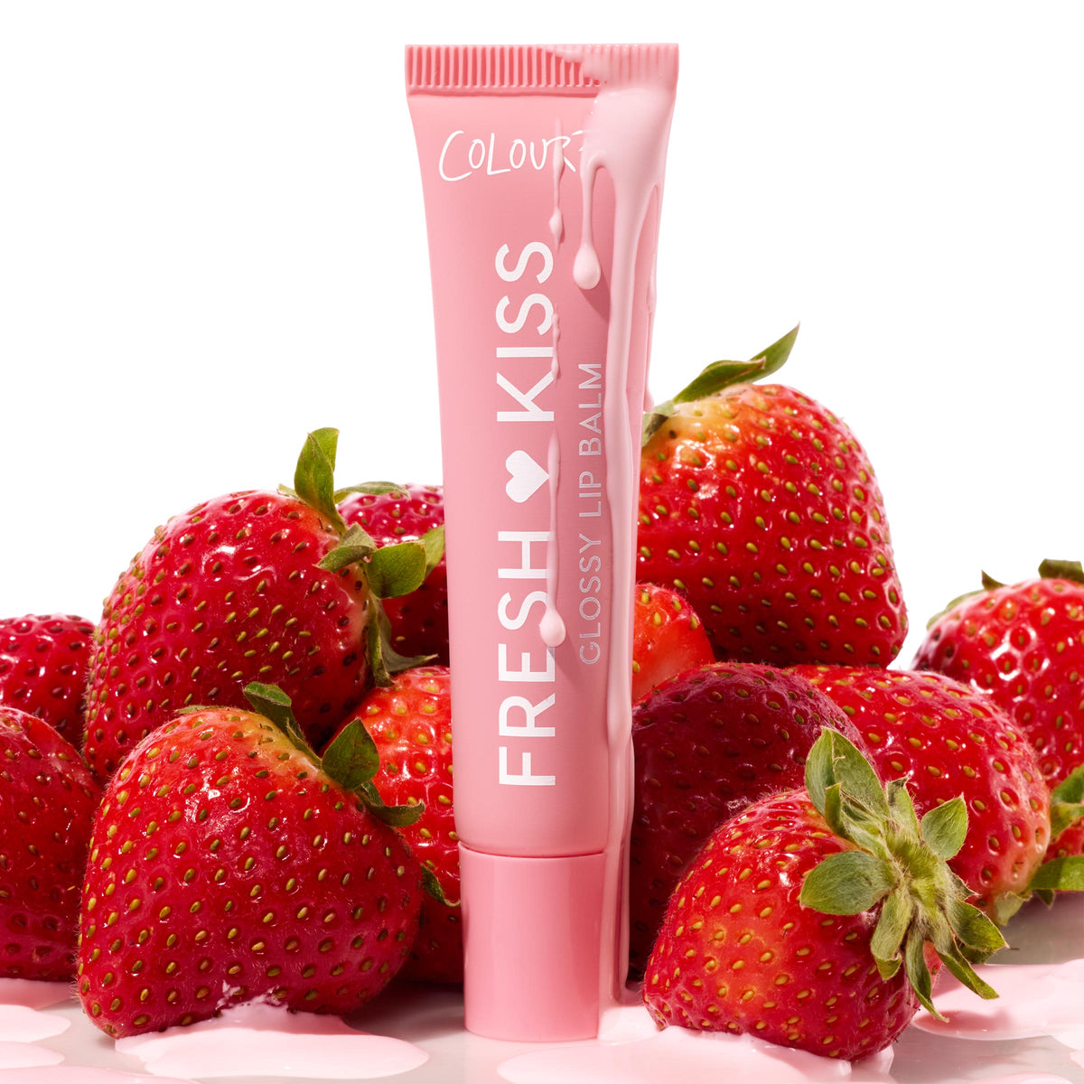 Pink ColourPop Fresh Kiss Glossy Lip Balm tube in Strawberry Milk stands upright, drizzled with pink cream, amid ripe strawberries on a white surface, highlighting Pretty in Pastels, the ColourPop Glossy Lip Balm Trio.
