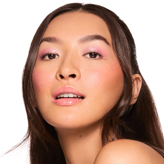 Model showcases glossy lips wearing ColourPop Fresh Kiss Glossy Balm in Watermelon Sugar, a sheer pink high-shine tint; close-up studio portrait with dewy skin, soft pink eyeshadow and blush against a white background.