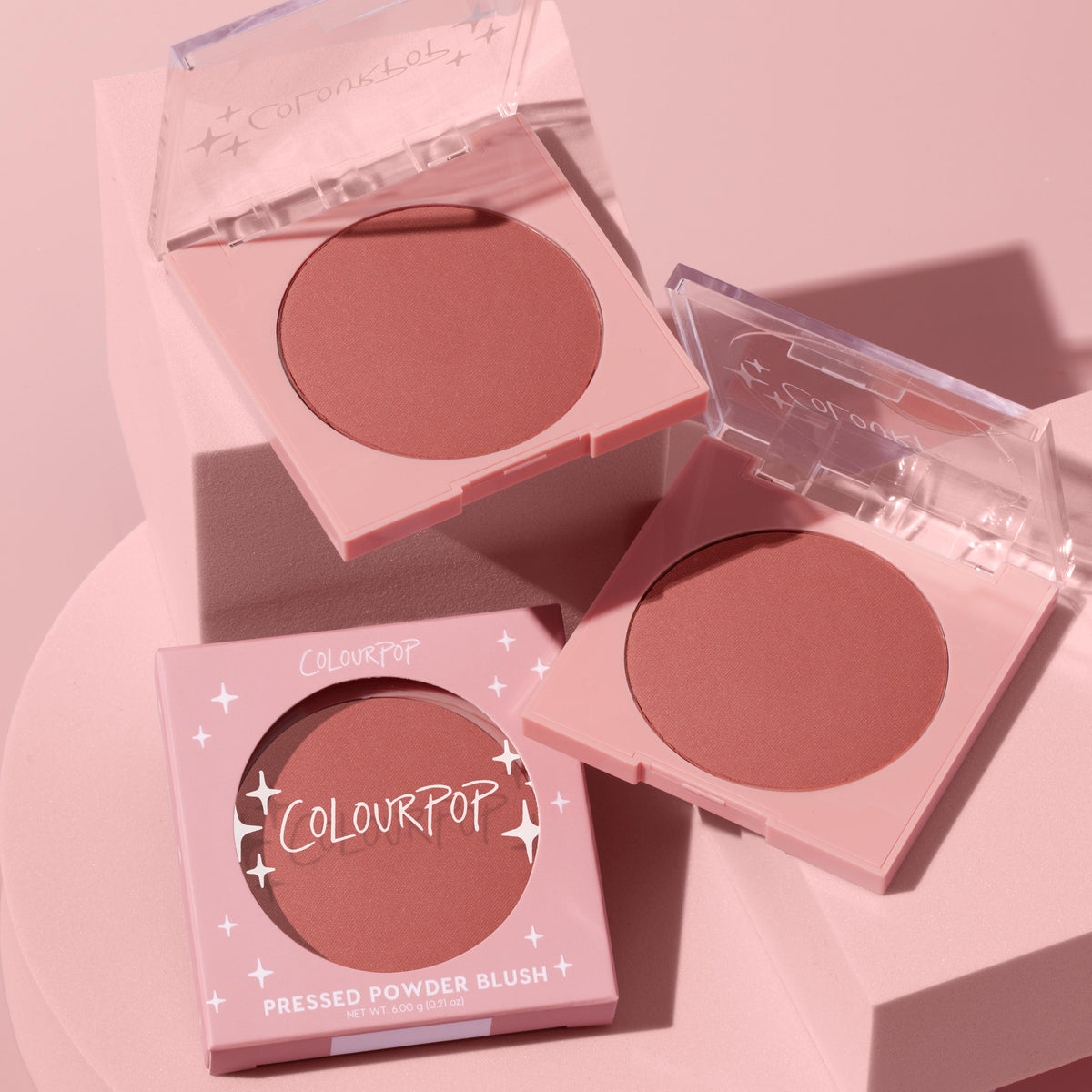 Just Cruisin’ ColourPop Pressed Powder Blush opens to reveal a warm rose shade in a pink compact with a clear lid, beside a second open pan and the retail box, styled on blush‑pink pedestals under soft studio lighting.