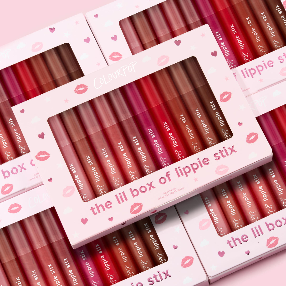 The Lil Box of Lippie Stix ColourPop Lippie Stix set displayed in windowed pink boxes, showcasing assorted nude, pink, and red shades; multiple kits stacked on a pastel pink background with hearts and kiss prints.