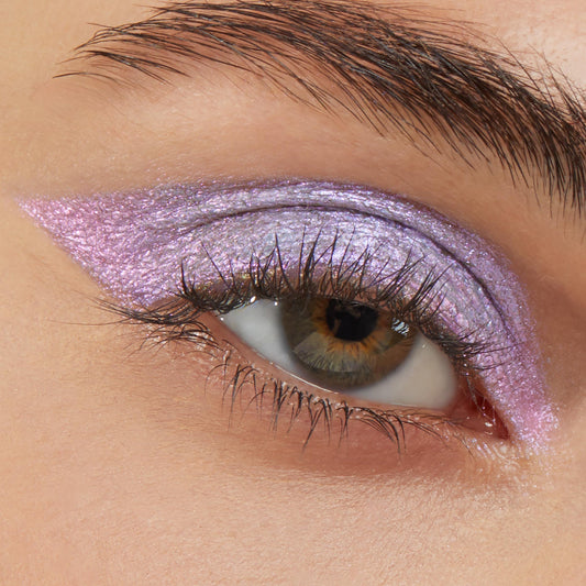 Close-up of a brown eye wearing Colourpop Cotton Candy Shadow Stix, showing a bold, winged wash of shimmering pastel lavender-pink cream eyeshadow across the lid and outer corner on smooth, natural skin.