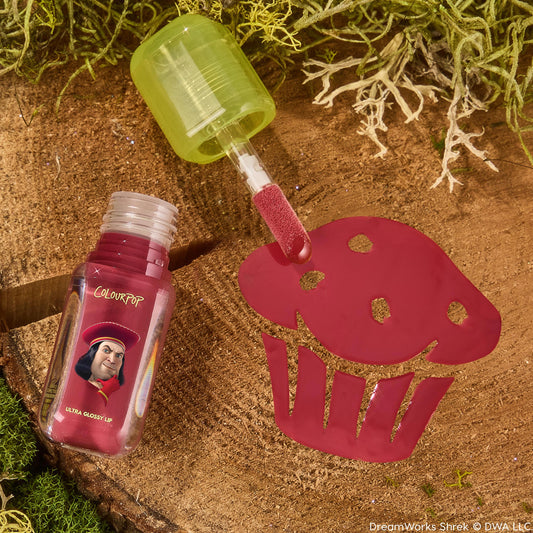 Short Lord Ultra Glossy Lip by ColourPop, applicator swatching raspberry-pink gloss through a cupcake stencil on a wood stump with moss; translucent green cap and bottle art featuring Lord Farquaad from Shrek; ColourPop Ultra Glossy Lip.