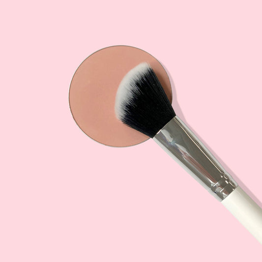 Angled Face Brush sweeping across a peach blush pan on a soft pink background, featuring black-and-white synthetic bristles, a silver ferrule, and a white handle, for contouring and blush application with ColourPop makeup.