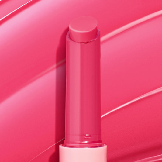 Tea Sesh gloss balm stick stands upright with the bright pink bullet extended and gleaming, set against sweeping hot-pink swatches; ColourPop So Juicy Plumping Gloss Balm with Peptides in a translucent pink tube with a light pink base.