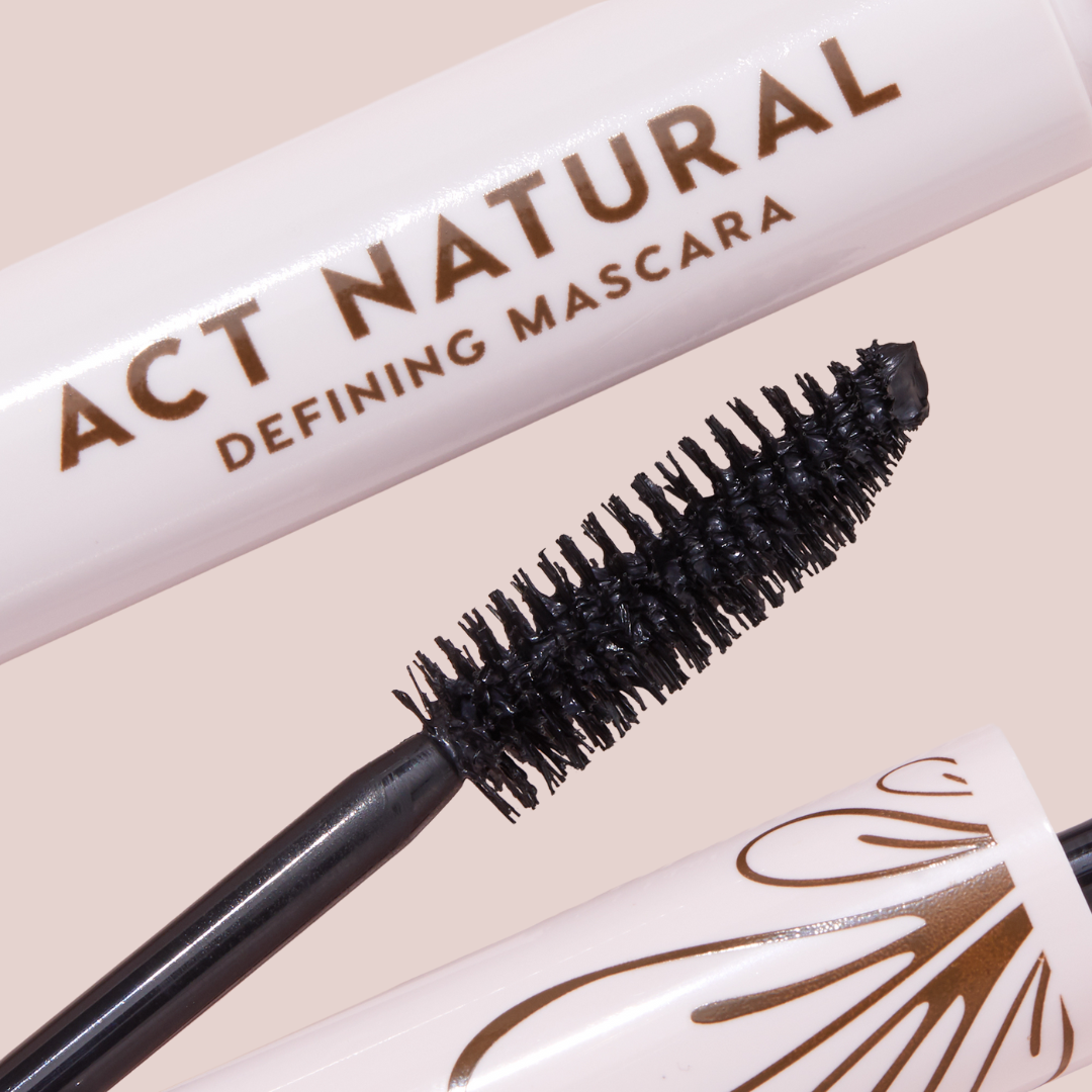 Mascara wand coated in deep black formula defining dense bristles, shown beside a blush-pink tube reading ACT NATURAL; ColourPop Act Natural Mascara, Black shade, with bronze detailing on the cap, close-up product shot on a neutral background.