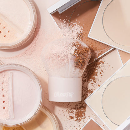 ColourPop Kabuki Face & Body Brush buffs loose and pressed powder, dense rounded bristles coated with pink and tan pigments, ColourPop Makeup Brush resting on a vanity amid open compacts and jars in light-to-tan shades.