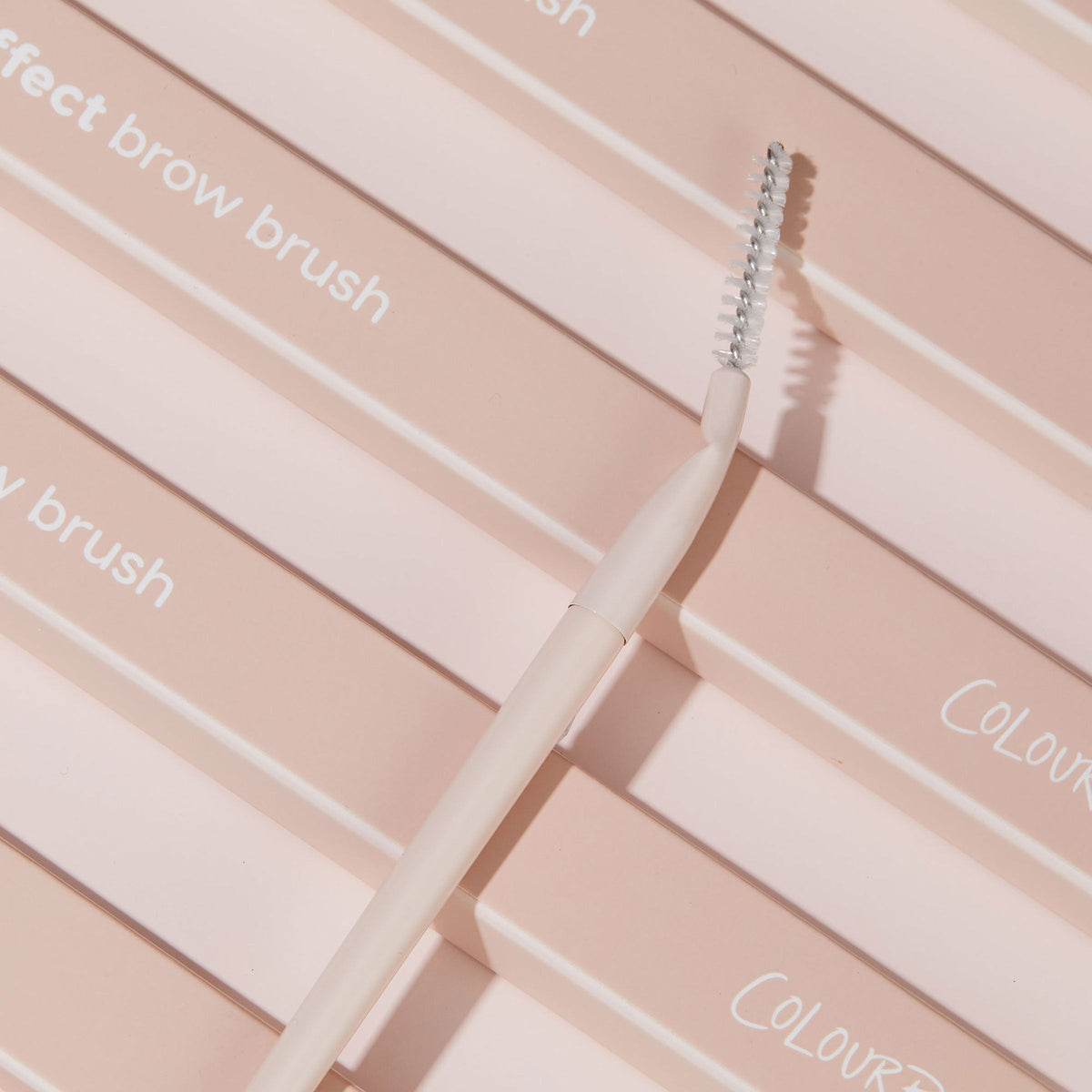Brow Brush spoolie angling across stacked blush-pink ColourPop boxes, featuring a slim cream handle, twisted neck, and silver spiral bristles; a makeup brush designed to shape and feather brows.