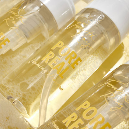 Clear pump bottles of Pore Real Cleanser foaming face wash glistening and tilted on a sudsy, golden surface, white caps and yellow lettering visible; ColourPop Face Cleanser by Fourth Ray Beauty highlighting pore-clearing, gentle lather.