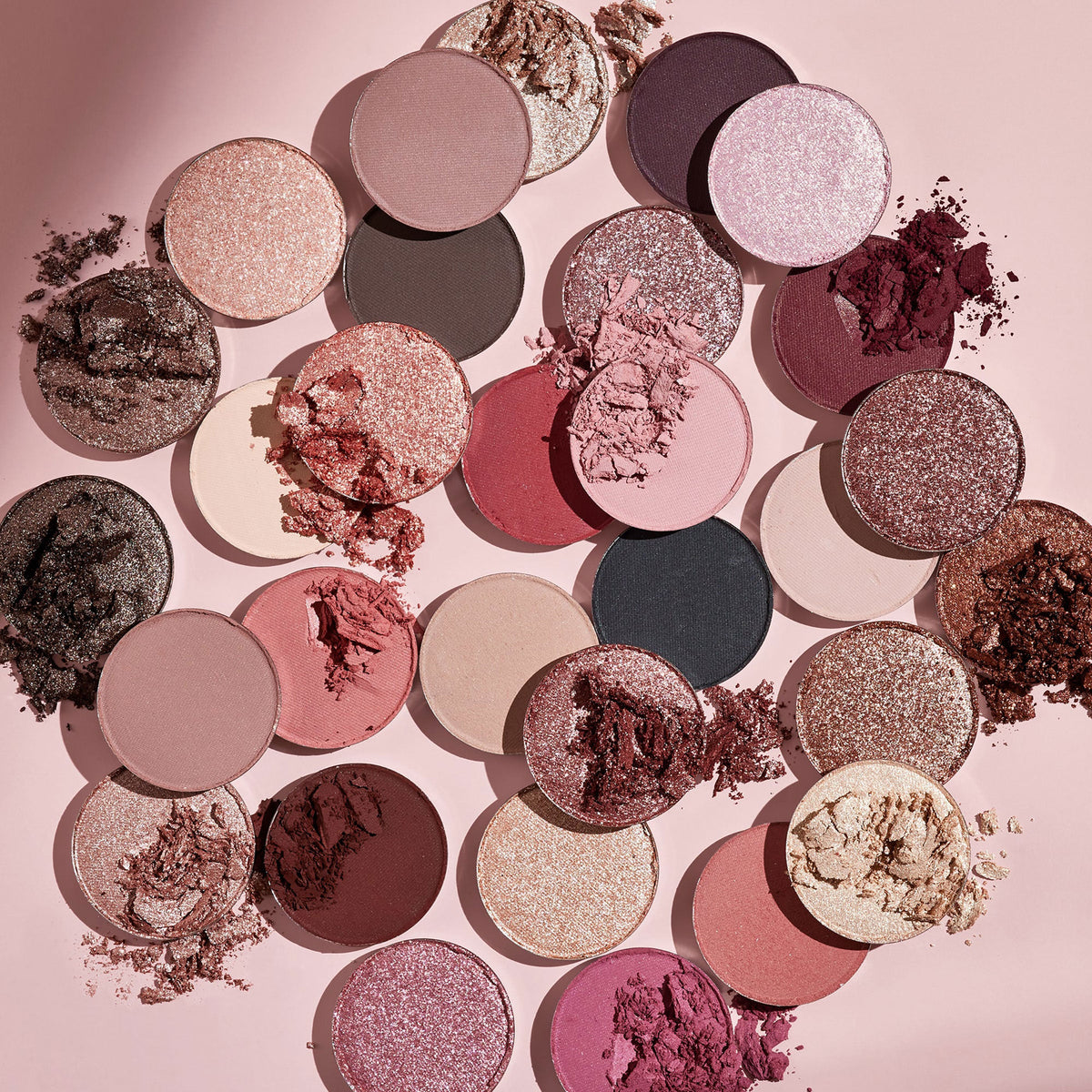 Smoke 'N Roses ColourPop Shadow Palette pans scattered on a pink background, showing round mattes and sparkly metallics in rose, mauve, plum, champagne, black, and nude tones with some crushed pigment for texture, ideal for romantic glam looks.