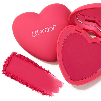 Heart-shaped ColourPop Pressed Powder Blush Text Me compact opens to reveal a vivid hot-pink pan, with a matching crushed swatch smeared beside it on a white background, showcasing matte fuchsia packaging with the ColourPop logo.
