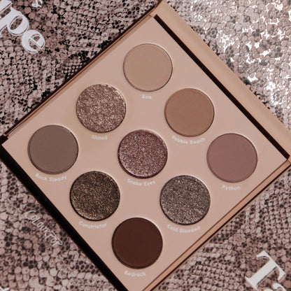 ColourPop Shadow Palette “That’s Taupe” open to nine cool‑toned taupe neutrals - mattes and metallic shimmers like Rock Steady, Slated, Snake Eyes, and Bedrock - arranged 3x3 on a beige tray atop a snakeskin-patterned background.