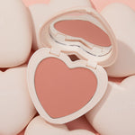ColourPop Kiss 'N Tell pressed powder blush in a heart-shaped compact, opened to reveal a matte peachy‑pink pan and mirror, sitting among cream heart compacts on a soft pink backdrop.