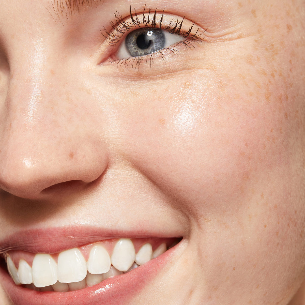 Lashes coated with ColourPop Act Natural Mascara in Brown, gently lengthening and separating a blue eye; close-up of a smiling face with freckles and dewy skin, showcasing a soft, natural brown mascara finish.
