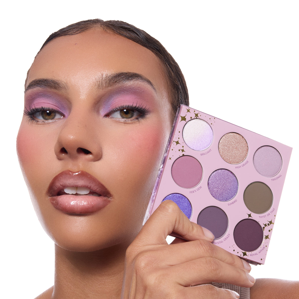 All Amethyst ColourPop Shadow Palette held beside a model’s lilac smokey eye, showing a 9‑pan lavender compact with gold star accents and matte-to-shimmer shades from soft pink and lavender to deep plum.