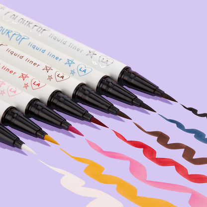 ColourPop Graceland BFF Liquid Liner creates a bold white squiggle beside multicolor swatches, the felt‑tip pen and doodled white barrel angled on a lavender background, showcasing precise application and opaque, quick‑dry wear.