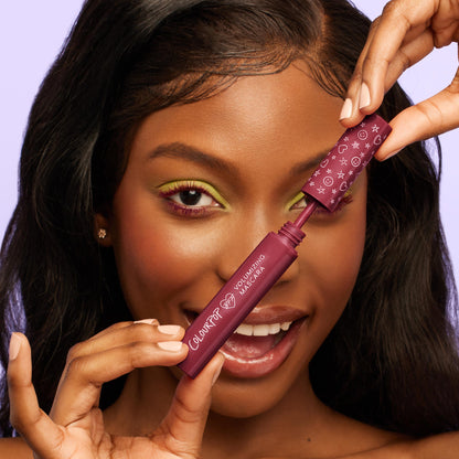 ColourPop BFF Mascara in Wine O’Clock, a burgundy volumizing formula, shown as a model opens the star-patterned wand near her lashes; close-up with lime green eyeshadow against a soft lavender background.