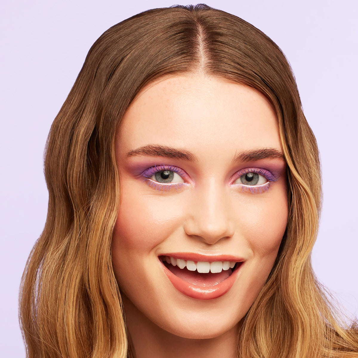 Lashes coated in ColourPop BFF Mascara in Purple Prose, fanning upward with bold purple pigment on upper and lower lashes, paired with lilac eyeshadow and glossy nude lips on a smiling model in a close-up against a pastel lavender background.