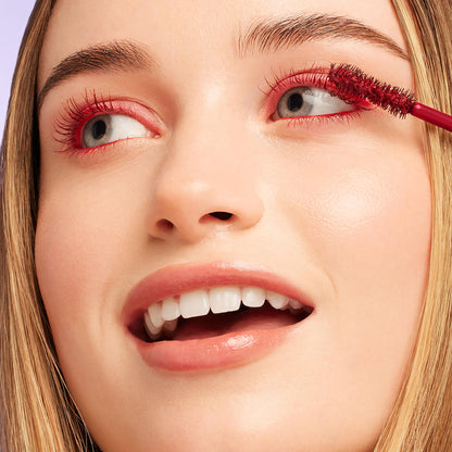 Mascara wand coats upper lashes in bold red, applying ColourPop BFF Mascara in Left on Red on a smiling model's eye, close-up studio shot with clean skin and neutral lips.