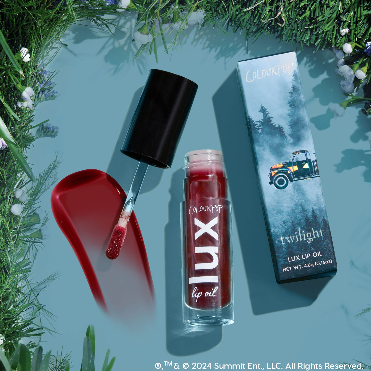 ColourPop Lux Lip Oil in shade Bells glides a glossy deep cherry swipe with a doe‑foot applicator, beside the clear LUX bottle and Twilight‑themed box with a pickup, on a teal flat lay framed by forest greenery.