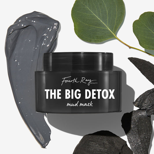 The Big Detox Mud Mask by Fourth Ray Beauty jar sits on a white backdrop beside a glossy gray mud smear, charcoal pieces, and green leaves, highlighting a detoxifying ColourPop Face Mask for deep pore cleansing.
