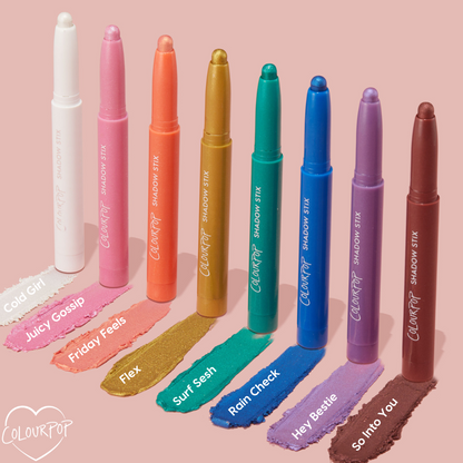 ColourPop Shadow Stix in Juicy Gossip - a shimmering bubblegum pink - shown with tip extended and a creamy swatch below, standing among other shades on a soft pink backdrop; twist-up eyeshadow crayon for quick, one-swipe color.