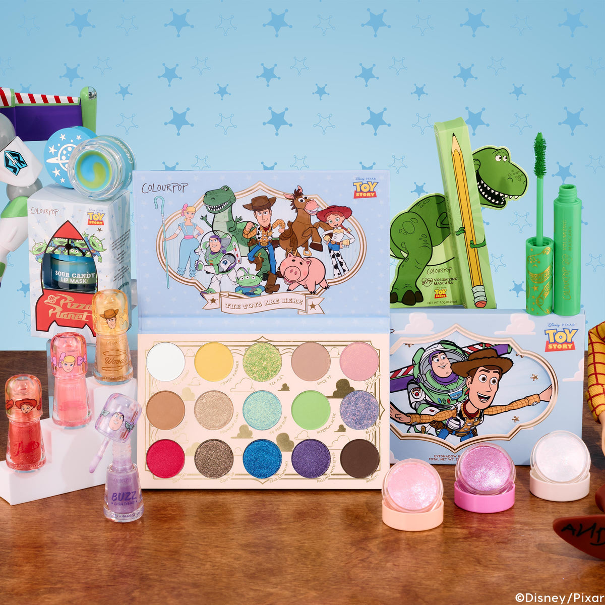 Toy Story and ColourPop Collection ColourPop Full Collection Set displayed on a wood table, showcasing a 15-shade eyeshadow palette, Rex green mascara, Buzz and Jessie lip glosses, shimmer pots, and a lip mask, against a starry blue Toy Story backdrop.