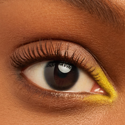 ColourPop Crème Gel Liner in Punch tracing a vivid yellow line along the inner corner and lower lash line of a brown eye, accenting natural lashes and neutral shimmer shadow in an extreme close-up on warm brown skin.