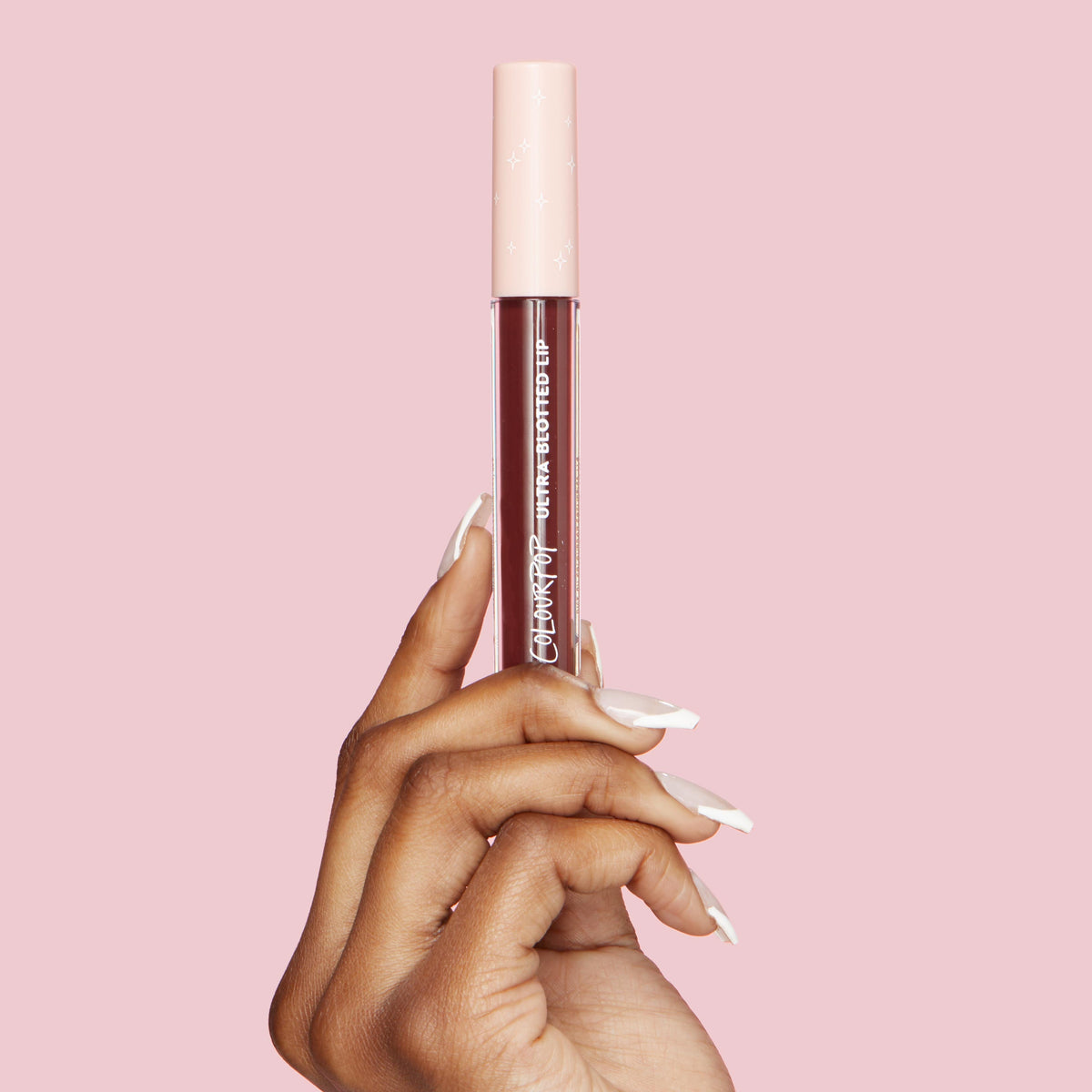 ColourPop Ultra Blotted Lip in Canon Dr held upright by a manicured hand, showcasing a clear tube of deep berry lip color with a pale pink star-print cap and white logo, set against a solid pink backdrop.