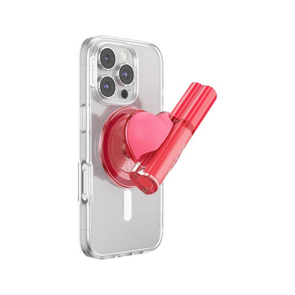ColourPopSockets ColourPop Lip Stain secured in a pink heart PopSockets grip on a clear smartphone case, the glossy pink tube angled outward against a white background, combining phone grip and lip stain holder for on‑the‑go use.