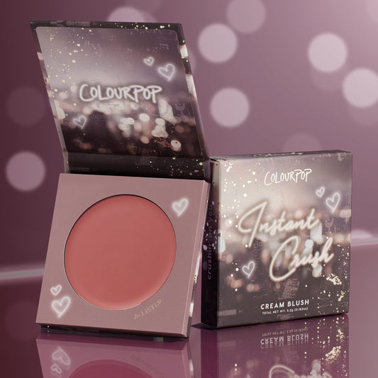 A-Lister cream blush compact sits open to reveal a warm rosy-mauve pan, with the matching box beside it; ColourPop Cream Blush Instant Crush packaging features heart doodles and sparkles against a mauve bokeh backdrop on a glossy surface.