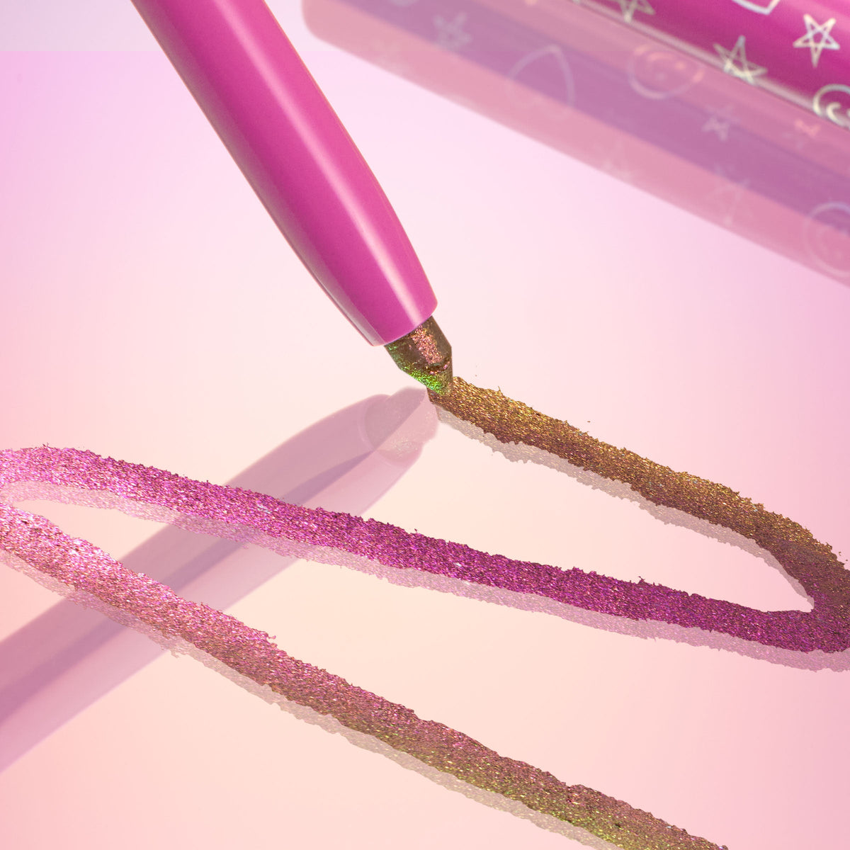 Pixie chrome crème gel liner gliding across a glossy pink surface, leaving duochrome strokes shifting from fuchsia to gold‑green, with the pink pencil barrel and star‑printed cap nearby; ColourPop Chrome Crème Gel Liner.