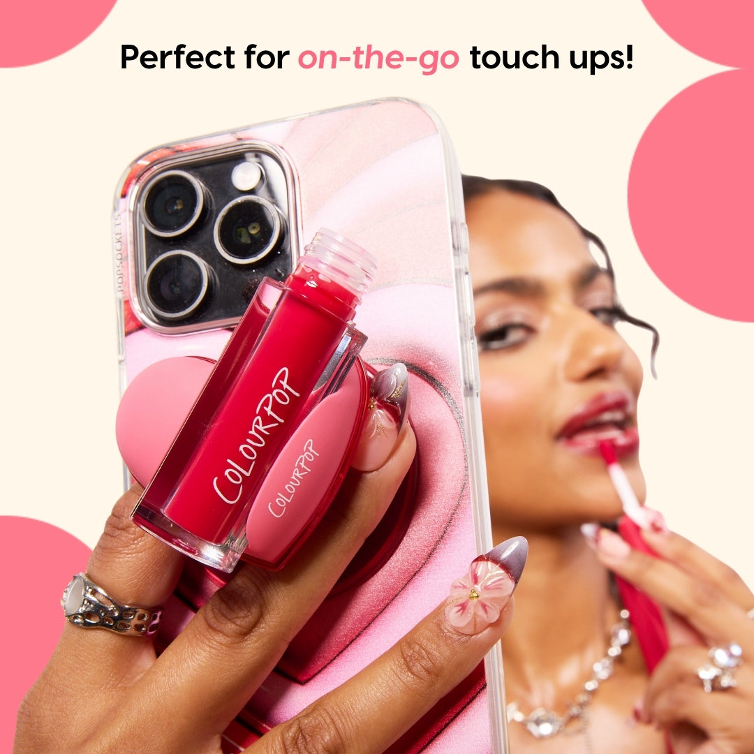 ColourPop Lip Stain in vivid pink, applied with a doe‑foot applicator, while a smartphone with a pink heart ColourPopSockets grip holds the slim rectangular tube for easy on‑the‑go touch‑ups; close‑up of floral nail art and glossy case.