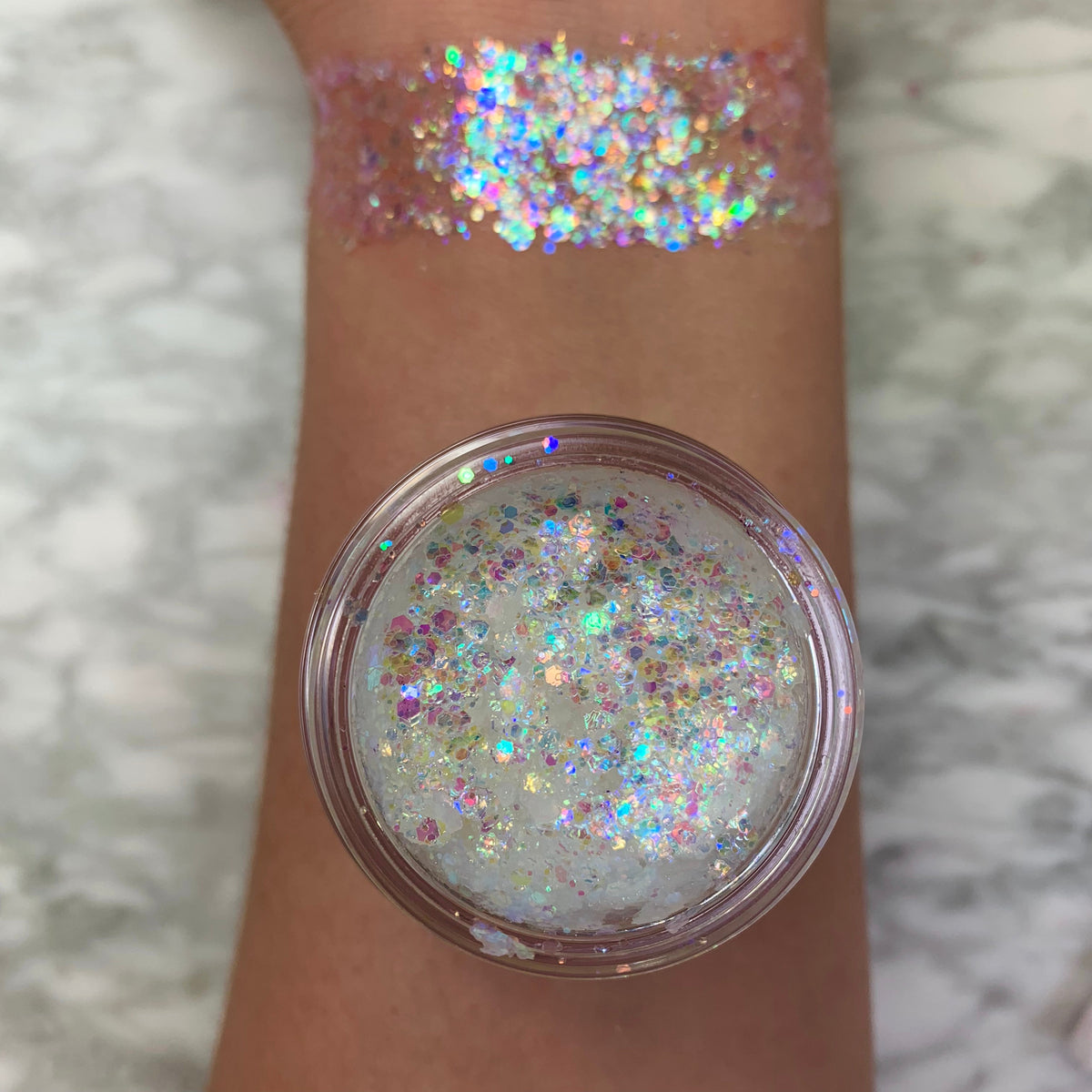 Disco Lady glitter gel swatched across a forearm, displaying chunky iridescent rainbow sparkle; an open pot of translucent gel packed with holographic flakes rests on the arm over a marble surface, showcasing payoff and texture; ColourPop Glitter Gel.