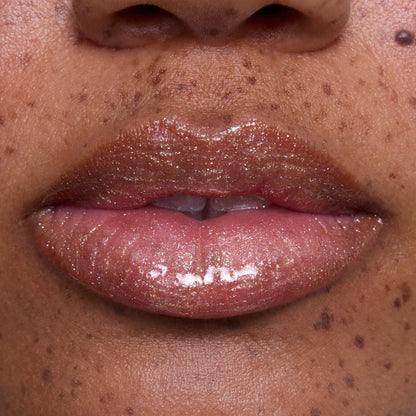 Close-up of glossy, medium-toned lips wearing Ambitious Tiana Ultra Glossy Lip, showing shimmering gold and pink sparkle inspired by Disney Princess beauty, with natural skin and freckles softly visible around the mouth.