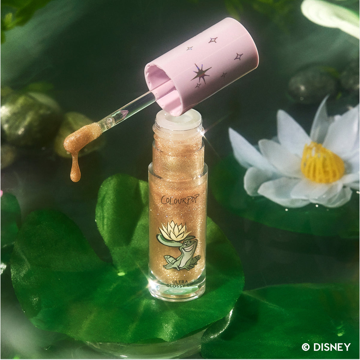 Ambitious Tiana Ultra Glossy Lip Disney Princess lip gloss with golden shimmer sits open on a green lily pad, wand lifted and dripping gloss, surrounded by water, lily pads, and a white lotus flower in a dreamy fairytale setting.