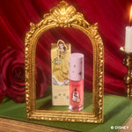 ColourPop Mademoiselle Belle Ultra Glossy Lip stands beside its yellow Disney Princess Belle box inside an ornate gold mirror frame, styled with a red rose, candle, and red velvet backdrop for a fairytale-inspired makeup product shot.