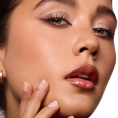 Glossed lips wearing ColourPop So Juicy Plumping Gloss Balm with Peptides, shade Perfection, delivering a plumped high-shine finish as a model touches her cheek; close-up beauty shot with neutral eyeshadow in studio lighting.
