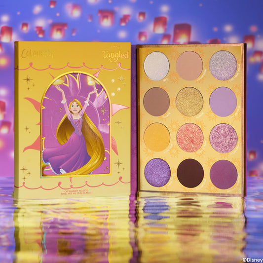 Best Day Ever ColourPop Shadow Palette shown open next to its Disney Tangled Rapunzel box, revealing a 12-pan mix of purple, gold, pink, and brown mattes and shimmers, set on reflective water with floating lanterns in the background.