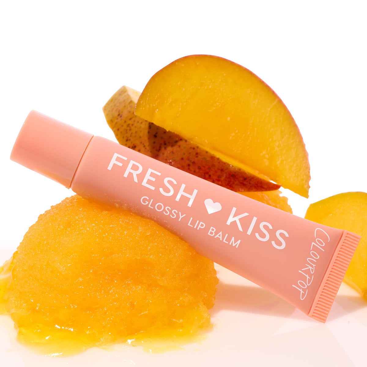 ColourPop Fresh Kiss glossy lip balm tube rests angled on a scoop of mango sorbet with mango slices, spotlighting the Pretty in Pastels ColourPop Glossy Lip Balm Trio; peach tube, hydrating shine on a clean white background.