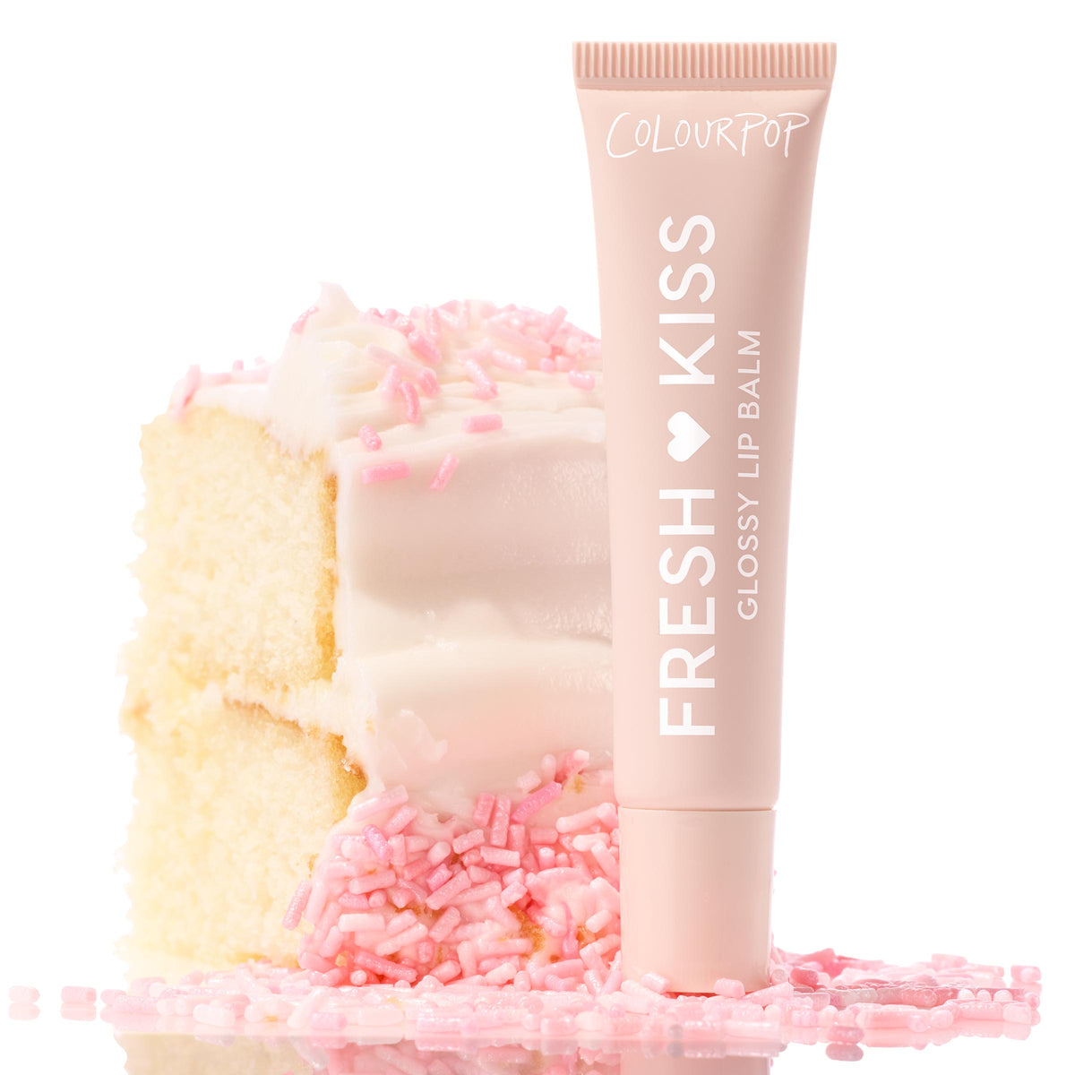 ColourPop Fresh Kiss Glossy Balm, Vanilla Cake, stands upright against a frosted vanilla cake slice topped with pink sprinkles, blush tube with white lettering styled on a bright, reflective surface for a sweet, high-shine lip finish.