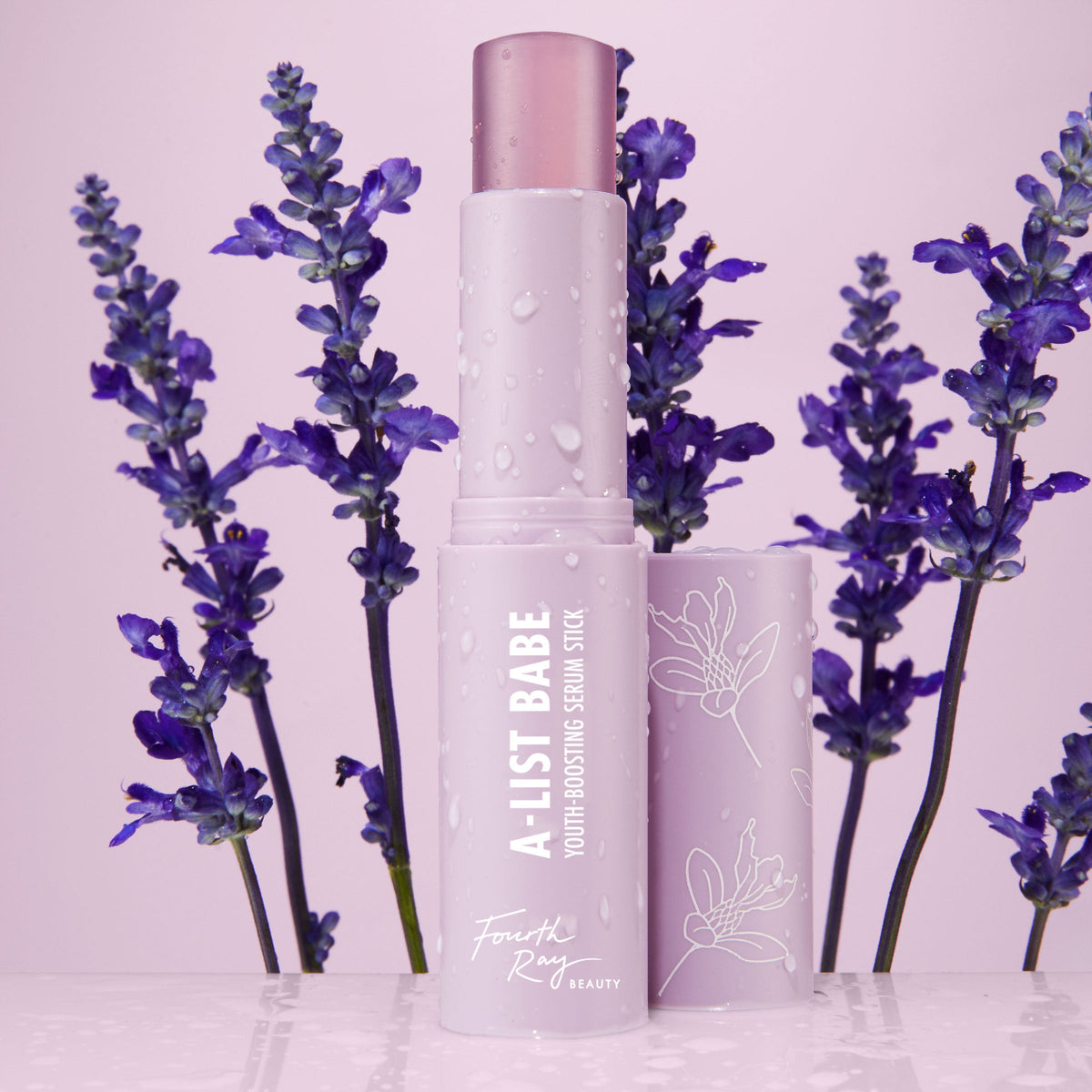 A-List Babe Serum Stick standing uncapped with dewy droplets on a pastel lavender tube, framed by purple lavender sprigs on a glossy pink surface; ColourPop Serum Stick by Fourth Ray Beauty for easy swipe-on face serum.