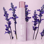 A-List Babe Serum Stick standing uncapped with dewy droplets on a pastel lavender tube, framed by purple lavender sprigs on a glossy pink surface; ColourPop Serum Stick by Fourth Ray Beauty for easy swipe-on face serum.
