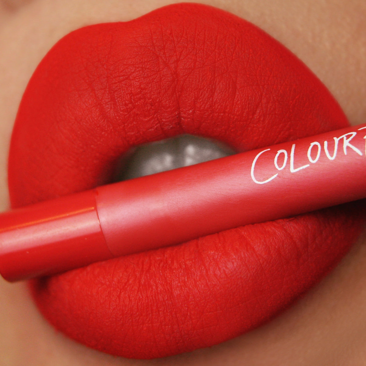 ColourPop Bring The Heat Lippie Pencil defining bold matte red lips, the vivid red pencil held between lips with the white ColourPop logo visible in a close-up, showcasing precise lining and vibrant, long-wear color.