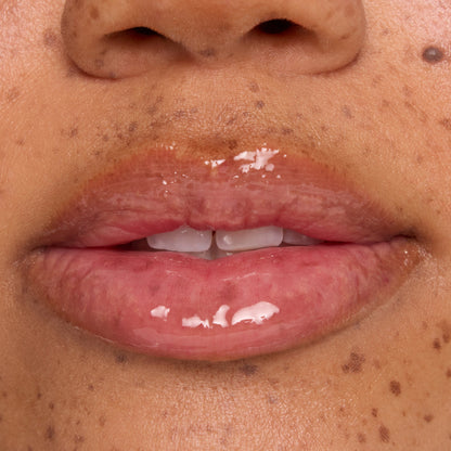 Glossy, hydrated lips wear a clear lip mask with small white balm flecks melting on the surface, shown in an extreme close-up on freckled skin to showcase ColourPop lip mask treatment shine and texture.