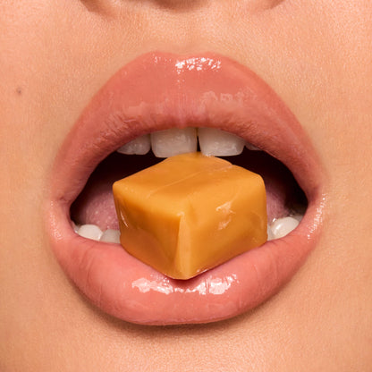 High-shine lips wear Caramel Latte, the ColourPop Fresh Kiss Glossy Balm, gripping a square caramel candy between the teeth in a tight macro close-up, highlighting the glossy finish, smooth texture, and warm nude sheen.