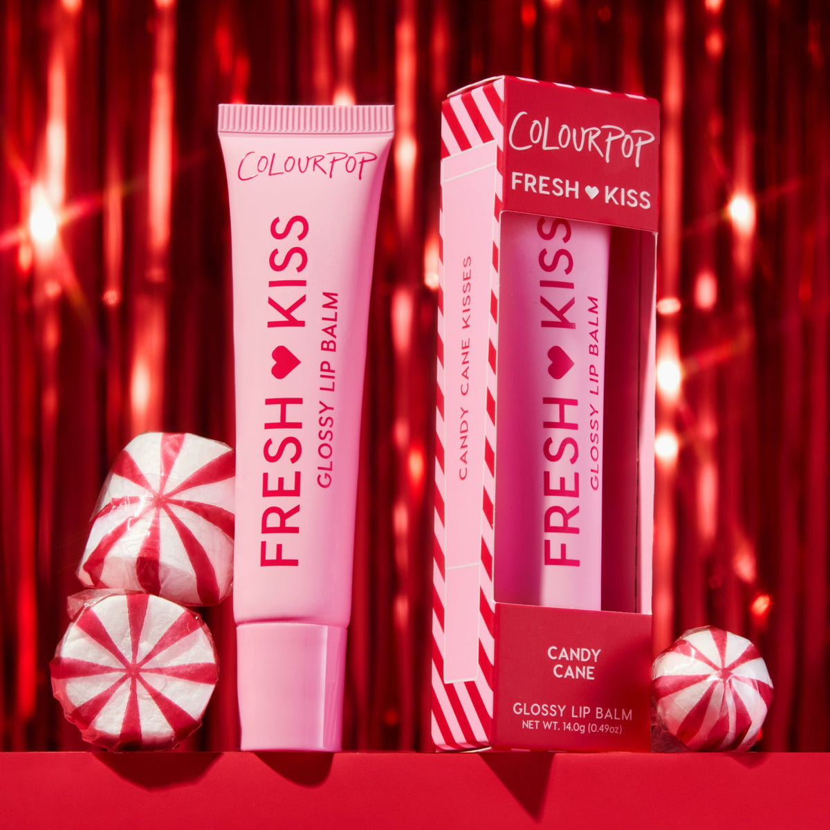 ColourPop Fresh Kiss Glossy Lip Balm, Candy Cane, stands upright beside its boxed twin, pink-and-red striped packaging, amid peppermint candies against a shimmering red tinsel holiday backdrop, 0.49 oz (14 g) tube.