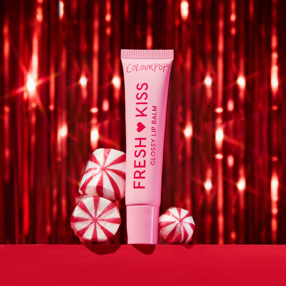 ColourPop Fresh Kiss Glossy Lip Balm in a pink squeeze tube stands upright beside red-and-white peppermint candies on a red surface, framed by sparkling red tinsel for a festive holiday beauty gift scene.